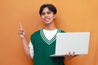 Yeşil kazak giyen neşeli genç Asyalı adam dizüstü bilgisayarını tutarken poz veriyor. Suratı asyalı adam dizüstü bilgisayarını turuncu arka planda tutarken poz veriyor.