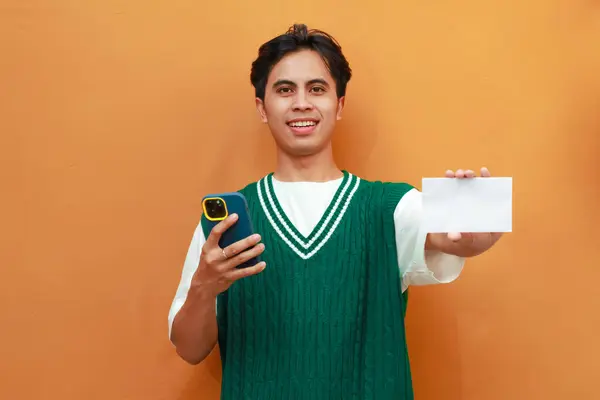 stock image Happy Young Asian man in green sweater posing holding mobile phone and showing blank white paper for advertisement at camera isolated on orange background