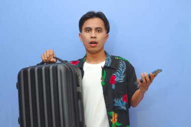 Plaj tişörtü giyen genç bir Asyalı bavul taşıyor ve şok olmuş bir ifadeyle cep telefonuna bakıyor.