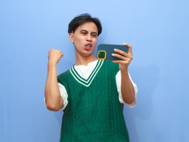 Akıllı telefondan mobil oyun oynayan Asyalı bir adamın portresi ya da canlı yayın videosu izleyen, gururla bağıran ve zaferi kutlayan ya da heyecanlı yüzüyle mavi arka planda kalan Asyalı bir adam..