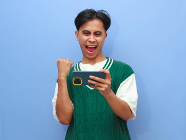 Akıllı telefondan mobil oyun oynayan Asyalı bir adamın portresi ya da canlı yayın videosu izleyen, gururla bağıran ve zaferi kutlayan ya da heyecanlı yüzüyle mavi arka planda kalan Asyalı bir adam..