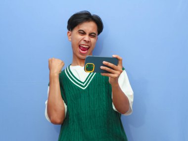 Akıllı telefondan mobil oyun oynayan Asyalı bir adamın portresi ya da canlı yayın videosu izleyen, gururla bağıran ve zaferi kutlayan ya da heyecanlı yüzüyle mavi arka planda kalan Asyalı bir adam..