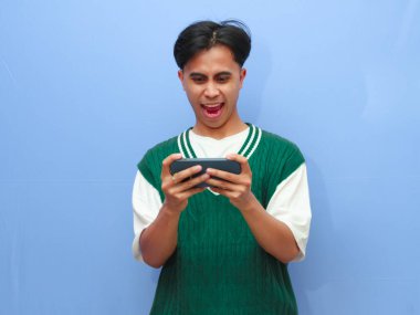 Akıllı telefondan mobil oyun oynayan Asyalı bir adamın portresi ya da canlı yayın videosu izleyen, gururla bağıran ve zaferi kutlayan ya da heyecanlı yüzüyle mavi arka planda kalan Asyalı bir adam..