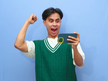 Akıllı telefondan mobil oyun oynayan Asyalı bir adamın portresi ya da canlı yayın videosu izleyen, gururla bağıran ve zaferi kutlayan ya da heyecanlı yüzüyle mavi arka planda kalan Asyalı bir adam..