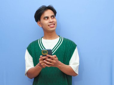 Cep telefonuyla poz veren sıradan giyinmiş mutlu Asyalı adam mavi arka planda..