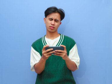 Akıllı telefon tutan Asyalı bir adamın portresi, izole edilmiş mavi arka planda stres jesti gösterirken mobil oyun oynuyor.