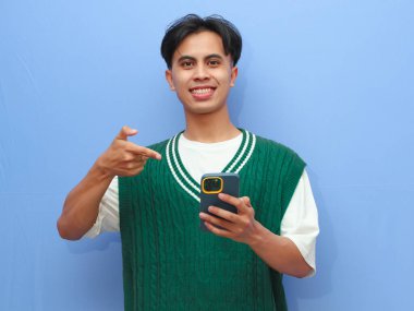 Cep telefonuyla poz veren sıradan giyinmiş mutlu Asyalı adam mavi arka planda..