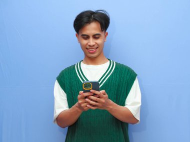 Cep telefonuyla poz veren sıradan giyinmiş mutlu Asyalı adam mavi arka planda..