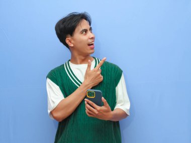 Cep telefonuyla poz veren sıradan giyinmiş mutlu Asyalı adam mavi arka planda..