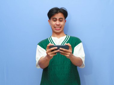 Asyalı bir adamın görüntülü video izlemesi veya mavi arka planda izole akıllı telefondan oyun oynaması komik bir surat ifadesi.