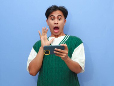 Asyalı bir adamın görüntülü video izlemesi veya mavi arka planda izole akıllı telefondan oyun oynaması komik bir surat ifadesi.