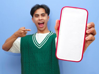 Asyalı bir adamın portresi beyaz ekranlı cep telefonu ve mutlu yüzü ile mavi arka planda izole edilmiş..