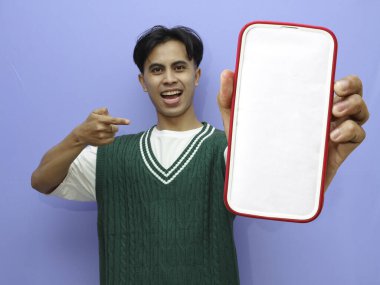 Asyalı bir adamın portresi beyaz ekranlı cep telefonu ve mutlu yüzü ile mavi arka planda izole edilmiş..