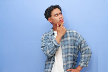 Genç Asyalı bir adam çenesini tutarken gözleri mavi arka planda ciddi bir yüzle düşünce kavramına bakıyor.