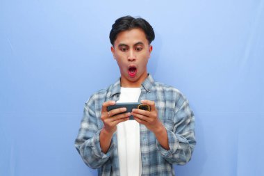 Mobil video oyunu uygulamasında akıllı telefon oyunu oynayan heyecanlı Asyalı genç oyuncu ya da mavi arka planda izole edilmiş dijital video akışı cep telefonunu izleyen