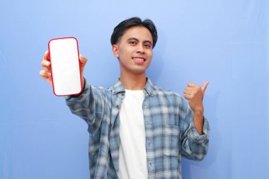 Mavi kareli gömlek giyen gülümseyen genç Asyalı adam bir reklam için cep telefonunu ekranda gösteriyor ve mavi arka planda izole edilmiş başarıyı kutluyor. Halk Yaşam Tarzı Konsepti