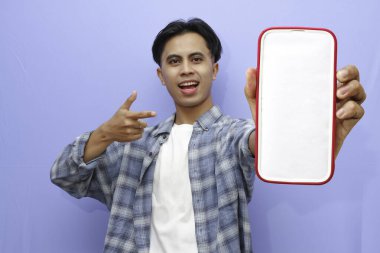 Mavi kareli gömlek giyen gülümseyen genç Asyalı adam bir reklam için cep telefonunu ekranda gösteriyor ve mavi arka planda izole edilmiş başarıyı kutluyor. Halk Yaşam Tarzı Konsepti