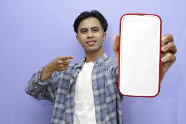 Mavi kareli gömlek giyen gülümseyen genç Asyalı adam bir reklam için cep telefonunu ekranda gösteriyor ve mavi arka planda izole edilmiş başarıyı kutluyor. Halk Yaşam Tarzı Konsepti