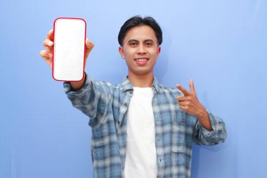 Mavi kareli gömlek giyen gülümseyen genç Asyalı adam bir reklam için cep telefonunu ekranda gösteriyor ve mavi arka planda izole edilmiş başarıyı kutluyor. Halk Yaşam Tarzı Konsepti
