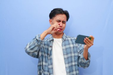 Üzgün surat Asyalı genç oyuncu mobil video oyunu uygulamasında akıllı telefon oyunu kullanıyor ya da mavi arkaplanda izole edilmiş dijital video akışı cep telefonunu izliyor