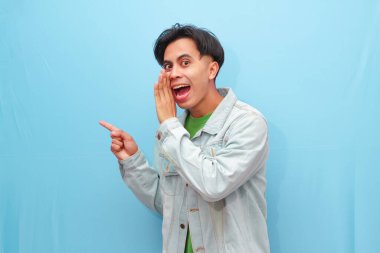 Asyalı genç bir adam fısıldayarak jest yapıyor. Heyecanlı bir yüzü var. Mavi arka planda.