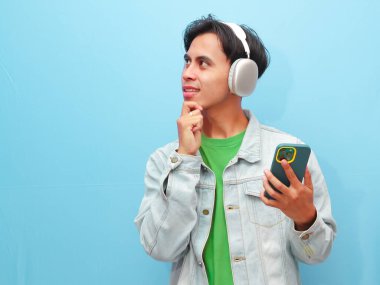 Yakışıklı Asyalı adam, kablosuz kulaklık takıyor ve akıllı telefon tutuyor. Düşünme hareketi yapıyor. Gözleriyle izole edilmiş mavi arka plana bakıyor.