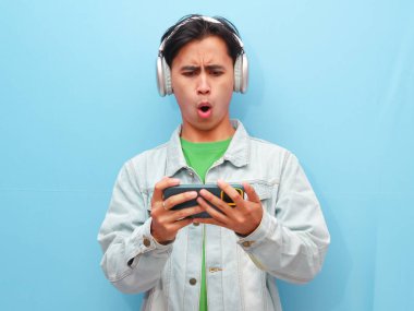 Mobil video oyunu uygulamasında akıllı telefon oyunu oynayan heyecanlı Asyalı genç oyuncu mavi arkaplanda izole edilmiş dijital video akışı cep telefonunu izliyor..