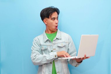 Asyalı genç adam elinde dizüstü bilgisayarla mutlu bir yüzle mavi arka planda poz veriyor.