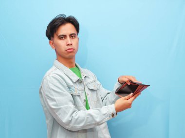 Zavallı ve bunalımlı Asyalı genç adam boş cüzdana bakarken mavi arka planda hüzünlü bir jest yapıyor..