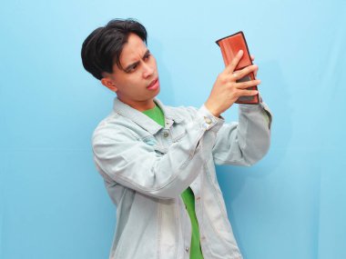 Zavallı ve bunalımlı Asyalı genç adam boş cüzdana bakarken mavi arka planda hüzünlü bir jest yapıyor..