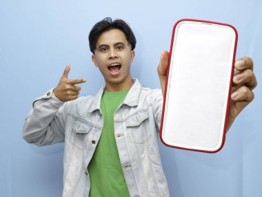 Mavi arka planda mutlu ve neşeli bir ifadeyle cep telefonuna poz veren sıradan giyinmiş genç bir Asyalı.