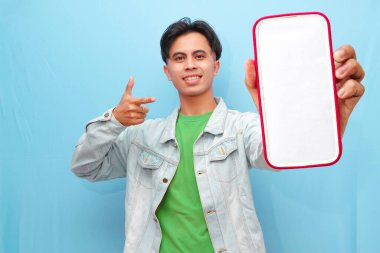 Mavi arka planda mutlu ve neşeli bir ifadeyle cep telefonuna poz veren sıradan giyinmiş genç bir Asyalı.