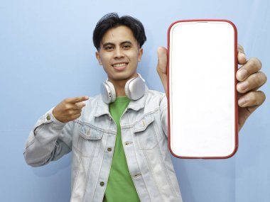 Mavi arka planda mutlu ve neşeli bir ifadeyle cep telefonuna poz veren sıradan giyinmiş genç bir Asyalı.