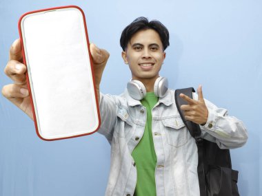 Sırt çantası taşıyan Asyalı bir erkek öğrenci. Elinde mutlu ve heyecanlı bir ifadeyle mavi arka planda cep telefonu taşıyor.