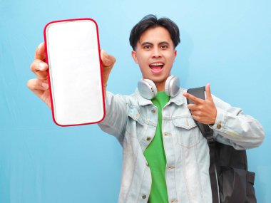 Sırt çantası taşıyan Asyalı bir erkek öğrenci. Elinde mutlu ve heyecanlı bir ifadeyle mavi arka planda cep telefonu taşıyor.