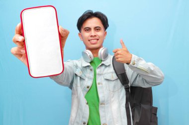 Sırt çantası taşıyan Asyalı bir erkek öğrenci. Elinde mutlu ve heyecanlı bir ifadeyle mavi arka planda cep telefonu taşıyor.