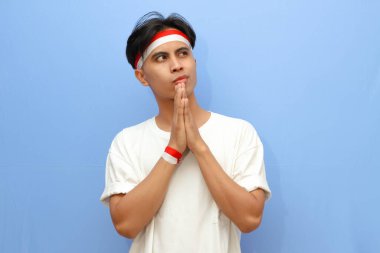 Beyaz tişörtlü Endonezyalı adam ve Endonezya bayrağı. Düşünme hareketi yapıyor. Çenesini ve gözlerini kaldırıp bakıyor..