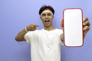Beyaz tişörtlü Endonezyalı adam ve Endonezya bayrağı mutlu yüzlü boş beyaz telefon ekranını işaret ediyor Endonezya bağımsızlık günü konsepti