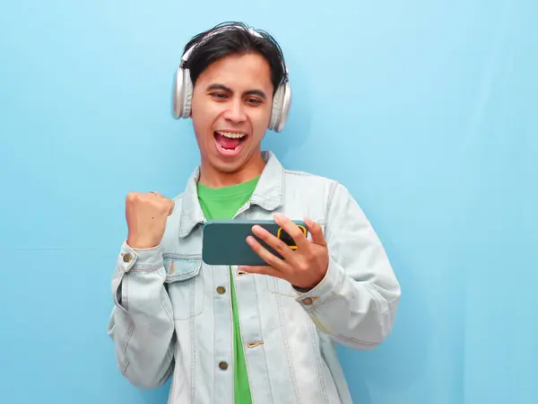 Mobil video oyunu uygulamasında akıllı telefon oyunu oynayan heyecanlı Asyalı genç oyuncu mavi arkaplanda izole edilmiş dijital video akışı cep telefonunu izliyor..