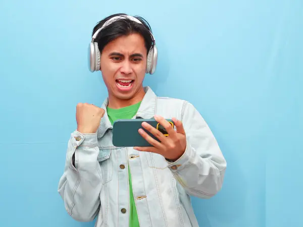 stock image Excited happy young Asian man gamer player using smartphone playing game in mobile video game app for watching digital video stream holding cell phone isolated on blue background.