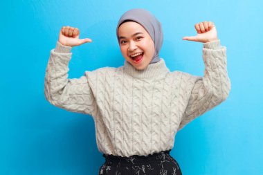 Neşeli, Asyalı, gri kazak giyen, yumruğunu sıkarak poz veren güzel Müslüman kadın, mavi arka planda mutlu bir jest yapıyor..