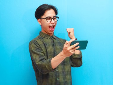 Mobil video oyunu uygulamasında akıllı telefon oyunu oynayan heyecanlı Asyalı genç oyuncu ya da izole edilmiş mavi arka planda dijital video akışını izleyen