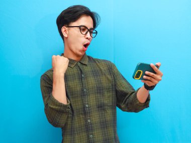 Mobil video oyunu uygulamasında akıllı telefon oyunu oynayan heyecanlı Asyalı genç oyuncu ya da izole edilmiş mavi arka planda dijital video akışını izleyen