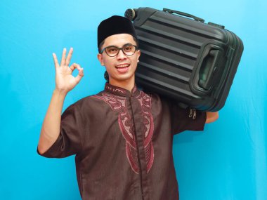 Portrait of excited indonesian muslim man carrying suitcase showing OK or Approved sign with hand  with smiling happy expression. Going home for Eid Mubarak or Mudik Isolated image on blue background. clipart