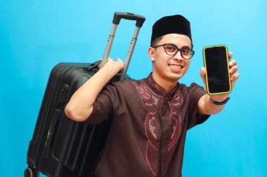 Portrait of a happy Indonesian Muslim man carrying a suitcase while holding a smartphone and showing a blank screen with excited gesture. Going home to celebrate Eid al-Fitr or Mudik Isolated image on a blue background. clipart