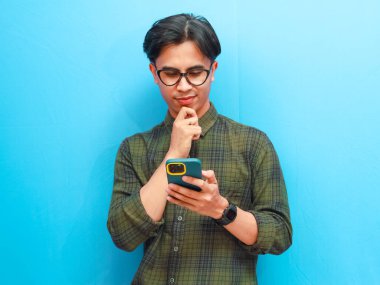 Resmi tişörtlü genç Asyalı adamın portresi. Akıllı telefon ticareti yapıyor ya da mavi izole stüdyo geçmişinde sohbet ediyor..