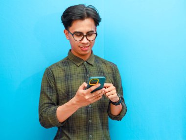 Resmi tişörtlü genç Asyalı adamın portresi. Akıllı telefon ticareti yapıyor ya da mavi izole stüdyo geçmişinde sohbet ediyor..