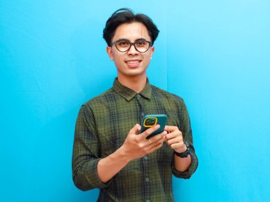 Resmi tişörtlü genç Asyalı adamın portresi. Akıllı telefon ticareti yapıyor ya da mavi izole stüdyo geçmişinde sohbet ediyor..