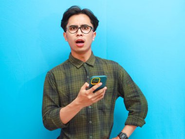 Elinde cep telefonu olan çekici Asyalı adam mavi arka planda şok olmuş bir ifade gösteriyor.