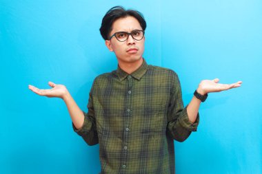 Yakışıklı Asyalı adam mavi arka planda ileriye bakarken şaşırmış ya da kafası karışmış bir ifade sergiliyor